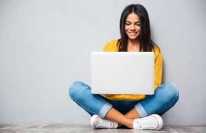 Happy-young-woman-sitting-on-the-floor-with-crossed-legs-and-using-laptop-on-gray-background (1)